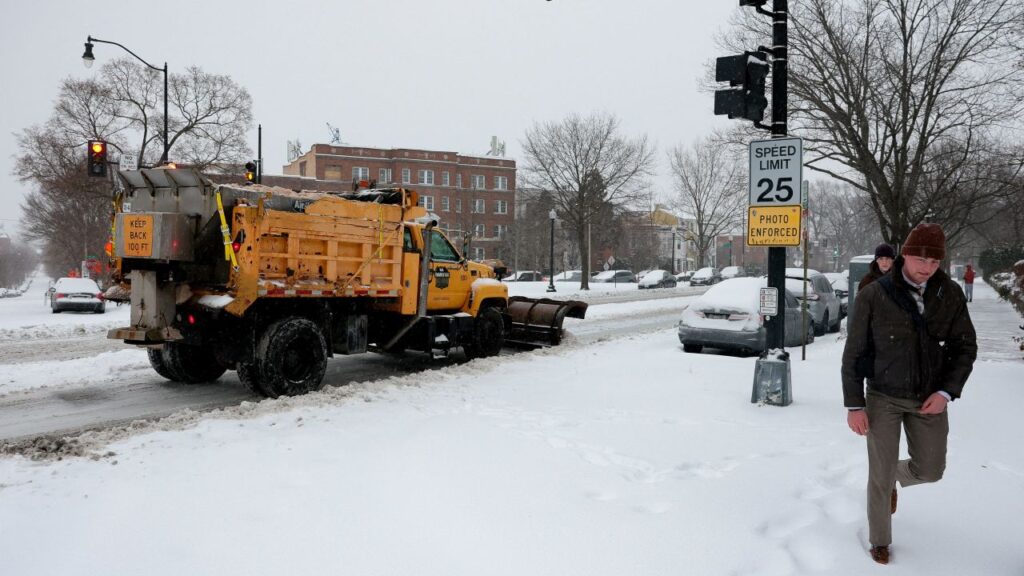 Image of a massive yellow snowplow clearing a street at a Washington, D.C. intersection. A man is walking nearby