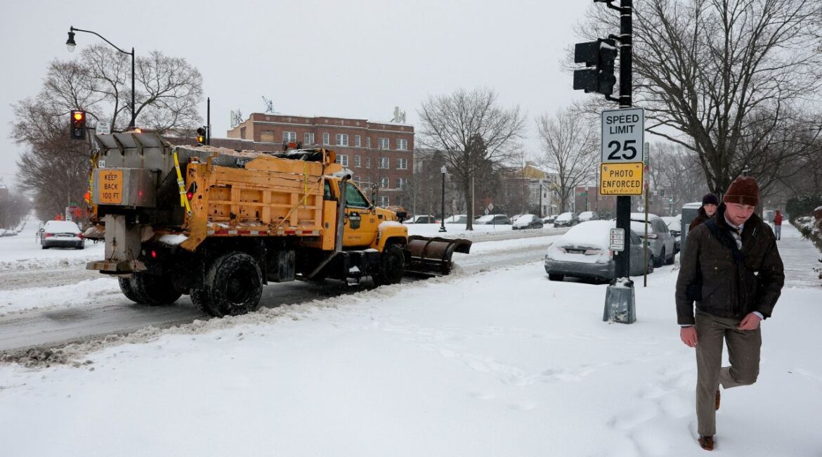 Image of a massive yellow snowplow clearing a street at a Washington, D.C. intersection. A man is walking nearby