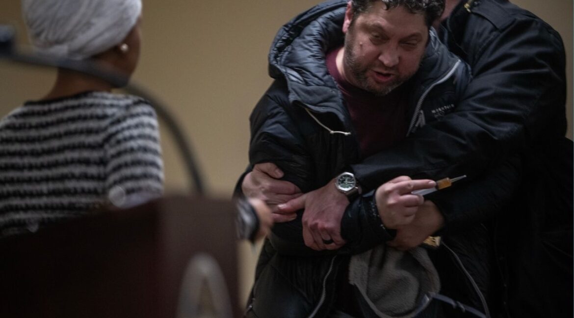 A man is held back after attacking Rep. Ilhan Omar (D-Minn.) during a town hall in Minneapolis, on Tuesday, Jan. 27, 2026. The man rushed the lectern and appeared to spray Omar with a strong-smelling liquid before he was tackled by security. Omar, visibly shaken after the assault, insisted to her staff and security that she would continue. (Victor J. Blue/The New York Times)