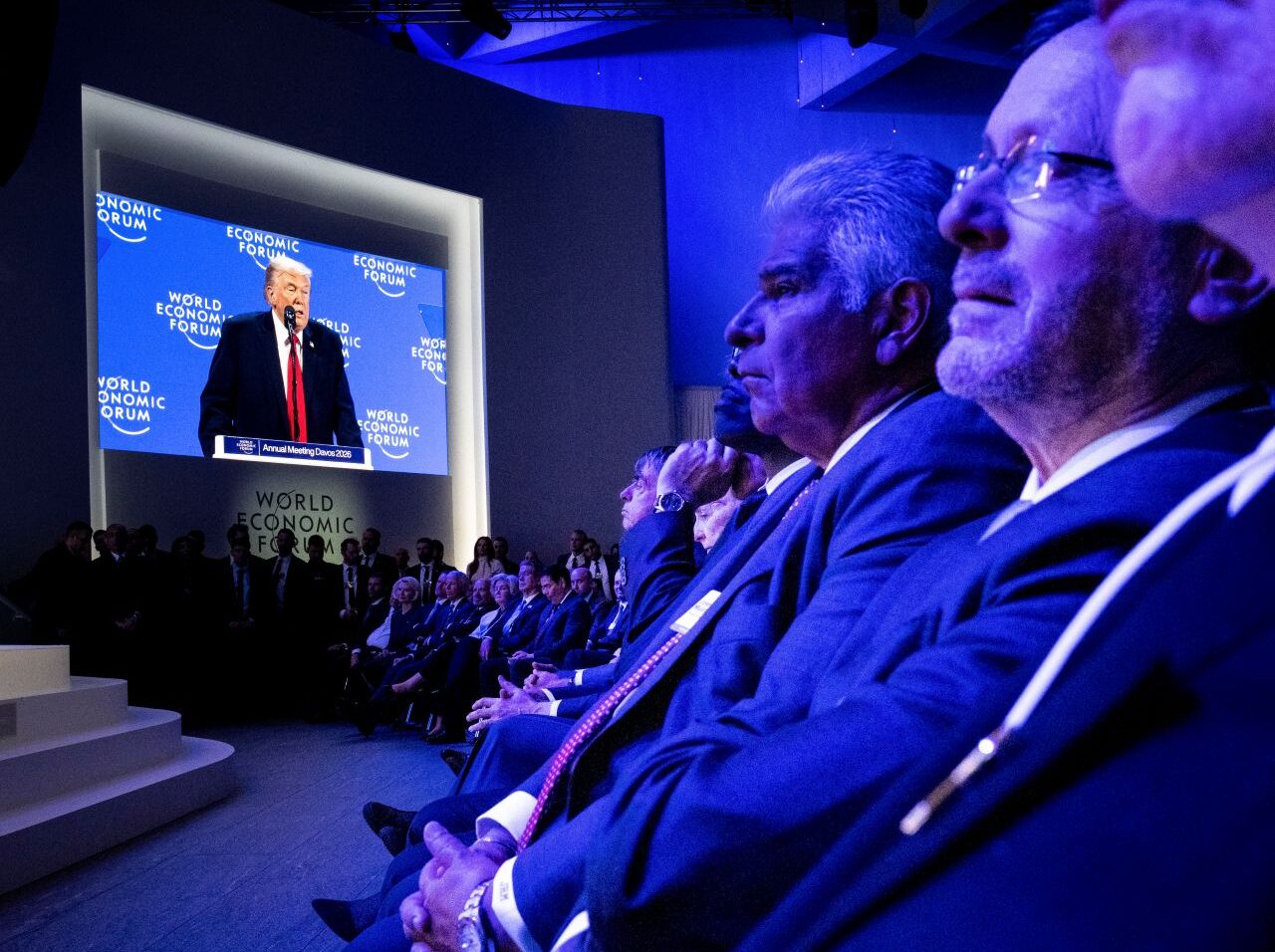 Image of members of the World Economic Forum viewing a speech by Donald Trump on a screen. The room is cast in a blue hue.