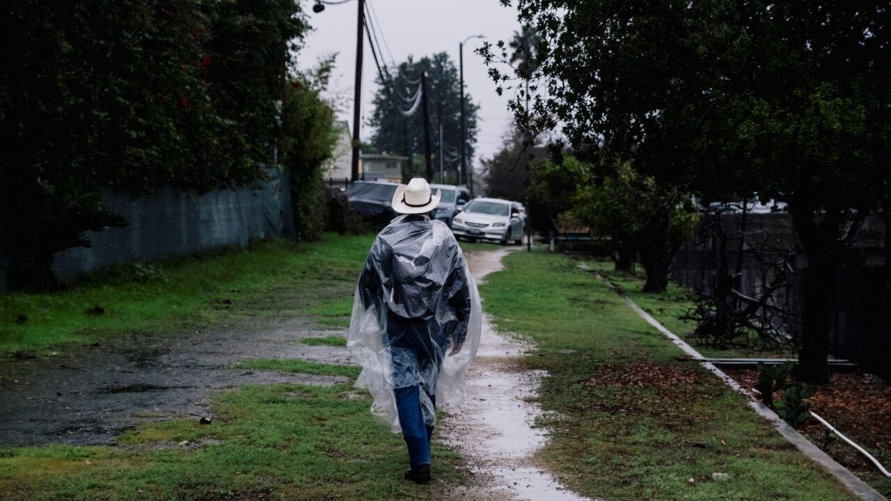 Image of a man walking down an alley. He's wearing a clear plastic poncho and a cowboy hat because of recent rain, which has puddled on the ground