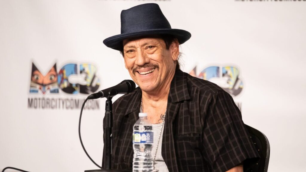 Image of the actor Danny Trejo speaking at a Comic Con event. He's wearing a black hat and coat.