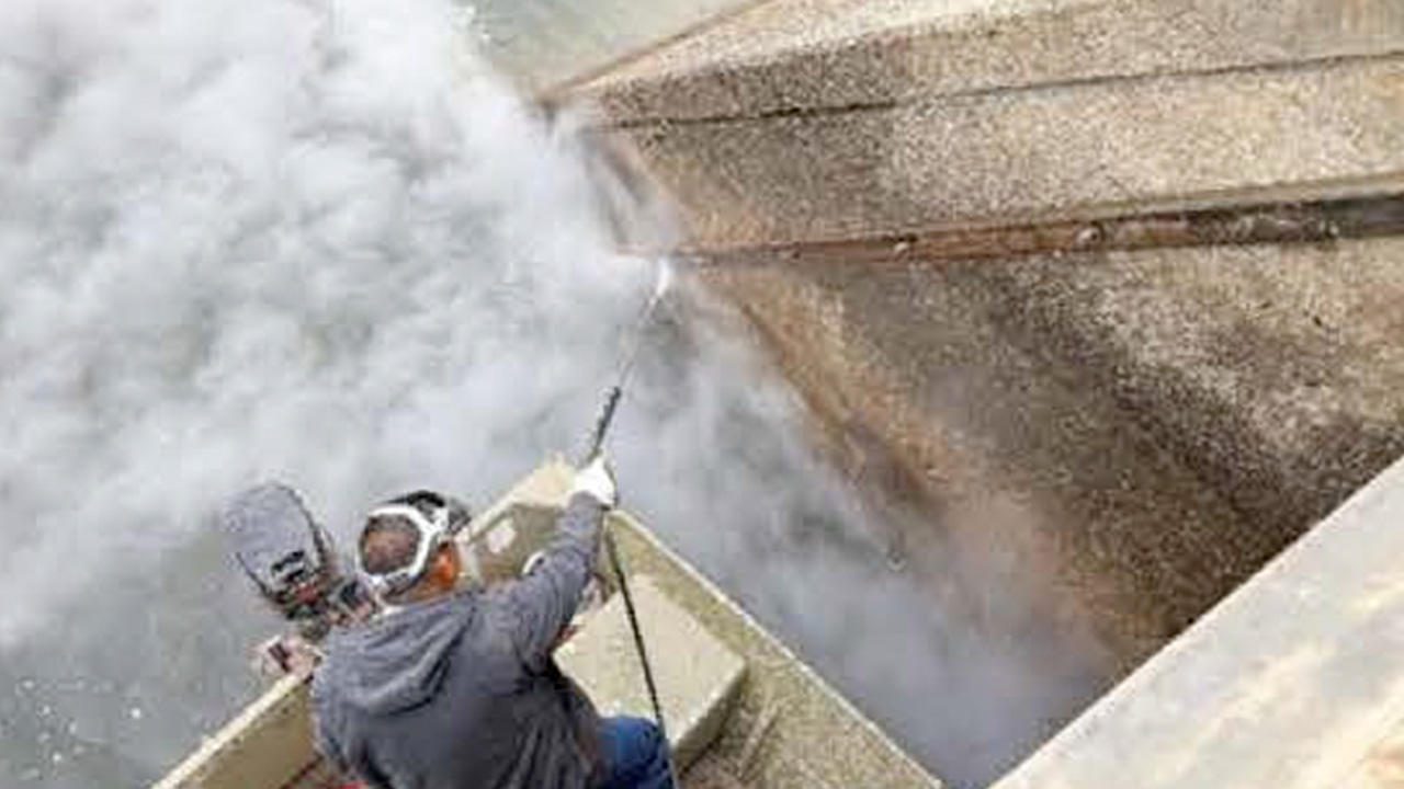 Image of a worker spraying super hot water on a concrete pillar that has golden mussels in the Friant-Kern Canal