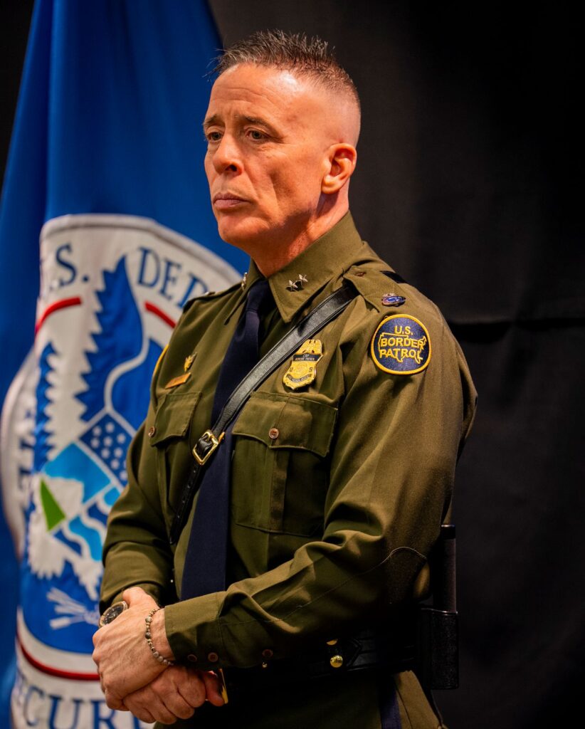 Image of Border Patrol Chief Gregory Bovino standing in front of a Homeland Security logo and blue curtain