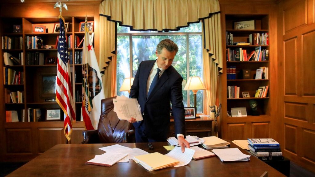 Image of Gavin Newsom in a dark suit and tie picking up papers from his governor's desk inside the California Capitol building