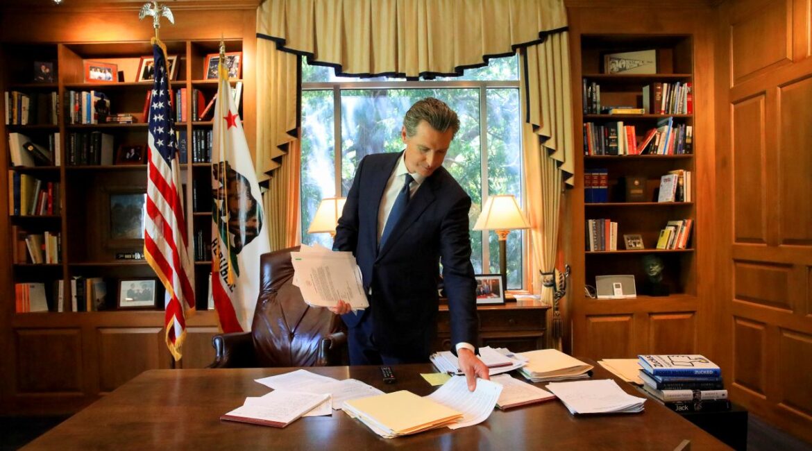 Image of Gavin Newsom in a dark suit and tie picking up papers from his governor's desk inside the California Capitol building