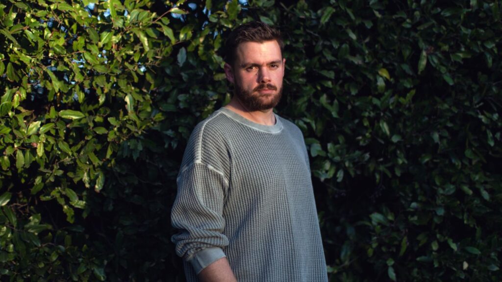 Image of Matt Schlicht standing among leafy shrubs and wearing a gray collarless shirt