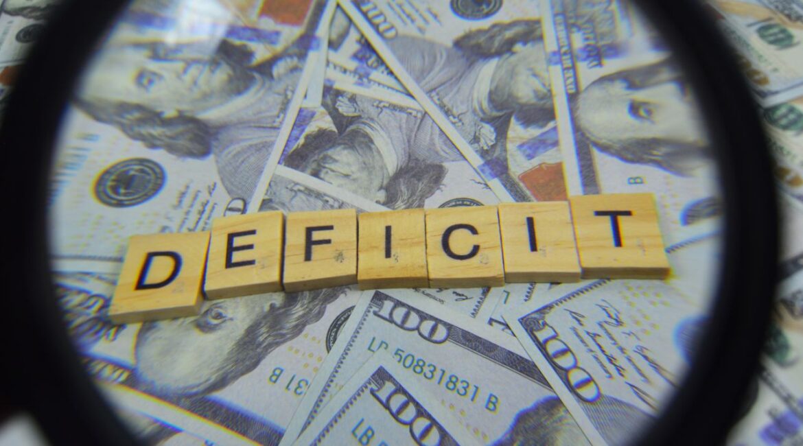 Scrabble tiles spell DEFICIT on a bed of $100 bills under a magnifying glass in this composite images