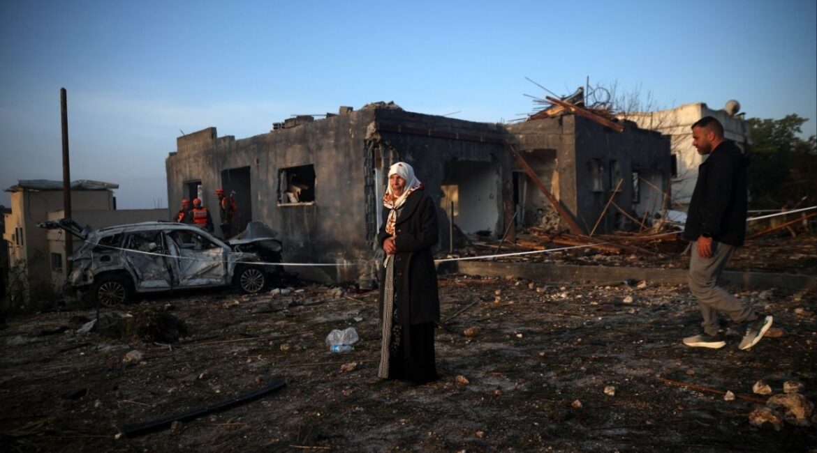 A woman stands as Israeli emergency responders work at the site of an impact by an Iranian missile, amid the U.S.-Israel conflict with Iran, in northern Israel, March 13, 2026. (Reuters/Shir Torem)
