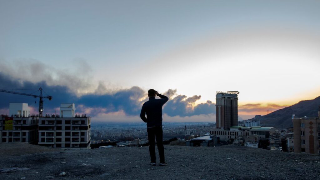Image of a man looking into the horizon at dusk as smoke rises from airstrikes in Tehran, Iran