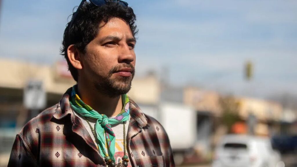 Image of Alfred Aldrete. He's wearing a wool shirt in a Native American pattern, has a beard, a green bandana and jewelry around his neck
