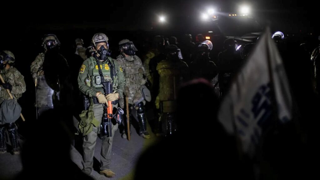 Image of federal immigration agents grouping up at night outside a farm. A few lights provide illumination