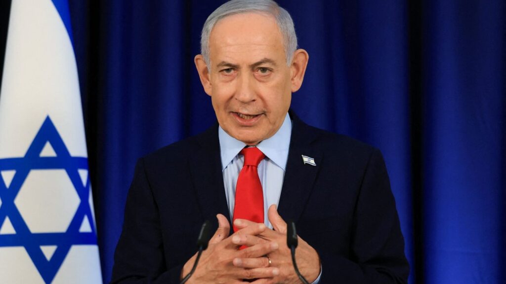 Image of Israeli PM Benjamin Netanyahu in a dark suit, blue shirt, and red tie. His hands are crossed and there is an Israeli flag to the side