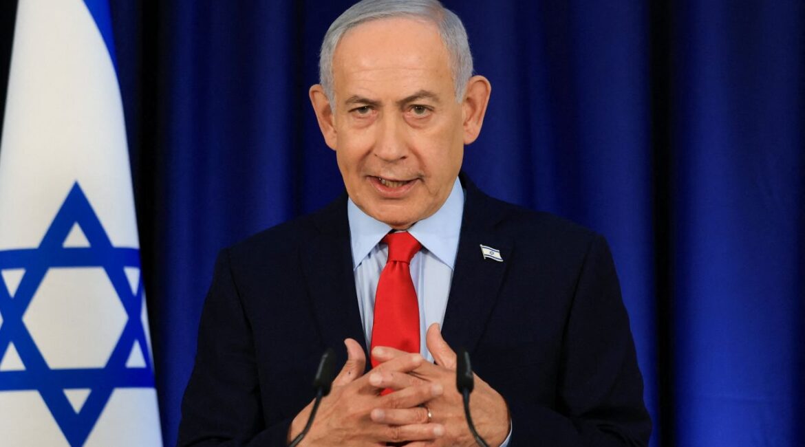 Image of Israeli PM Benjamin Netanyahu in a dark suit, blue shirt, and red tie. His hands are crossed and there is an Israeli flag to the side