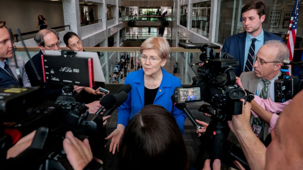 Image of Sen. Elizabeth Warren surrounded by media. She's wearing a black top and a light blue jacket.