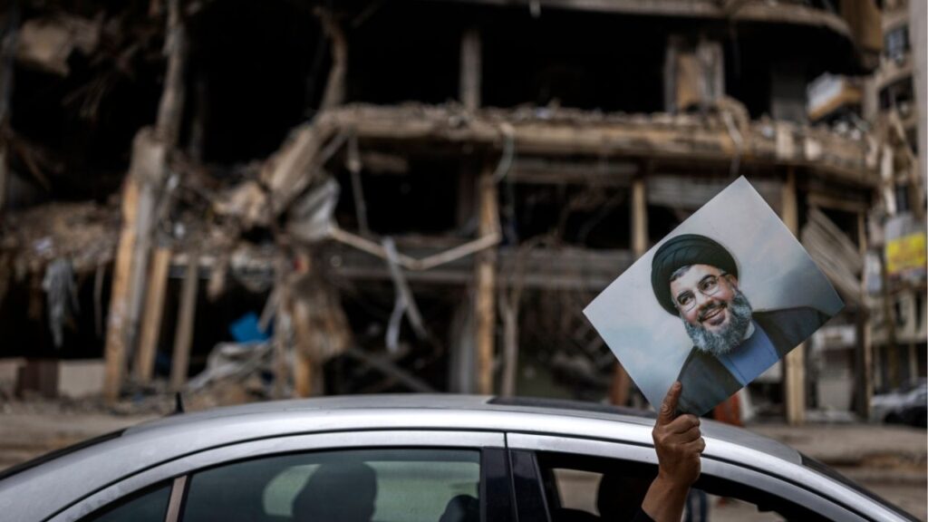 A man holds up a portrait of the slain Hezbollah leader Hassan Nasrallah as he is driven past the wreckage of a building destroyed in an Israeli airstrike in Dahiyeh, the suburb south of Beirut, Lebanon, on Monday, April 20, 2026. Joseph Aoun, the Lebanese president, said that he had appointed Simon Karam, a former ambassador to the United States, to lead talks with Israel aimed at ending the war and achieving a complete Israeli withdrawal from southern Lebanon. (Diego Ibarra Sánchez/The New York Times)