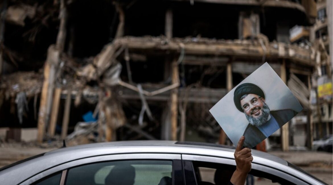 A man holds up a portrait of the slain Hezbollah leader Hassan Nasrallah as he is driven past the wreckage of a building destroyed in an Israeli airstrike in Dahiyeh, the suburb south of Beirut, Lebanon, on Monday, April 20, 2026. Joseph Aoun, the Lebanese president, said that he had appointed Simon Karam, a former ambassador to the United States, to lead talks with Israel aimed at ending the war and achieving a complete Israeli withdrawal from southern Lebanon. (Diego Ibarra Sánchez/The New York Times)