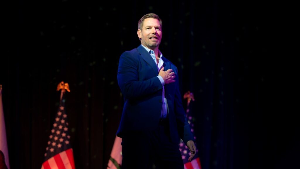 Image of Rep. Eric Swalwell standing on stage in a blue suit with an open collar shirt and his hand over his heart. There are American flags in the background.