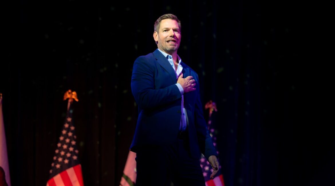 Image of Rep. Eric Swalwell standing on stage in a blue suit with an open collar shirt and his hand over his heart. There are American flags in the background.