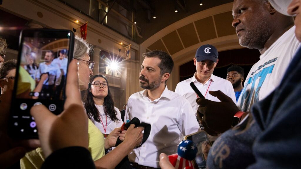 Image of Pete Buttigeg in a while shirt with an open collar surrounded by media