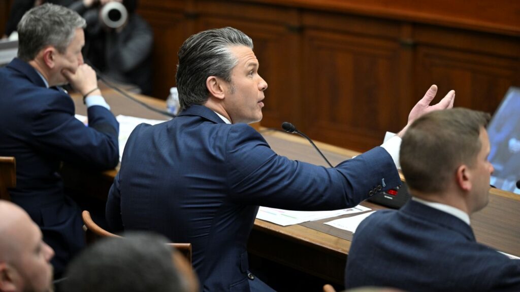 Image of Defense Secretary Pete Hegseth gesturing while speaking to the House Armed Services Committee. He's wearing a blue suit and white tie.