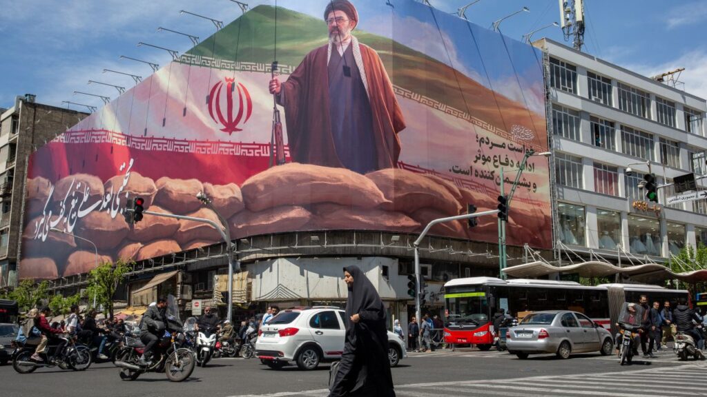Image of a woman walking across a square in Tehran and below a billboard depicting the new supreme leader.