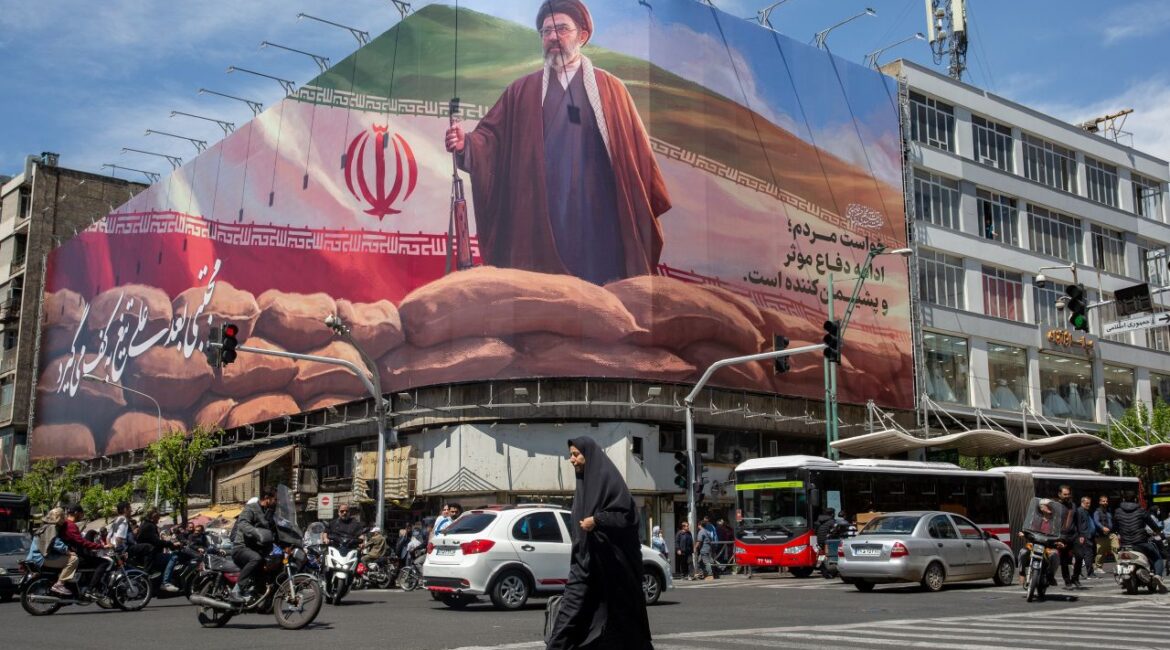Image of a woman walking across a square in Tehran and below a billboard depicting the new supreme leader.