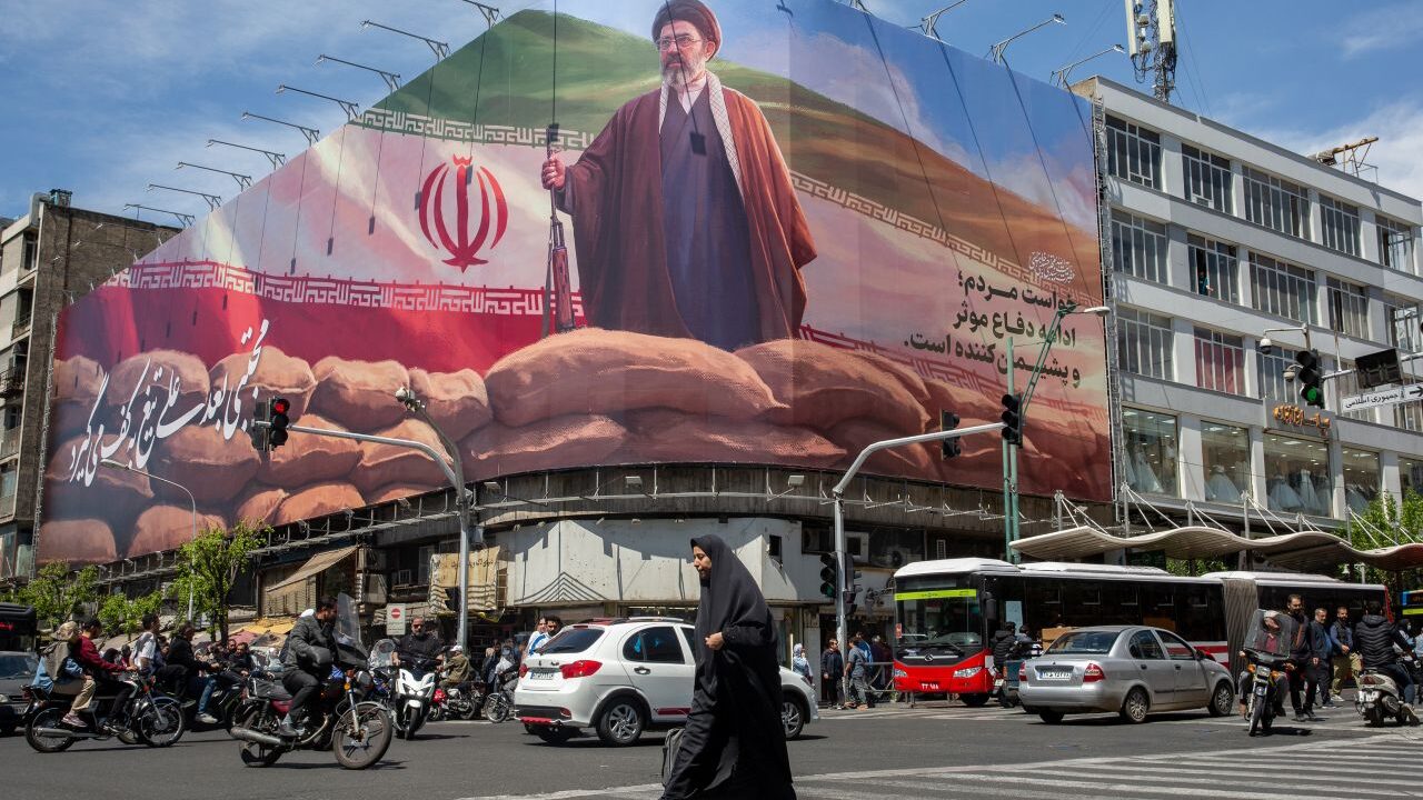 Image of a woman walking across a square in Tehran and below a billboard depicting the new supreme leader.