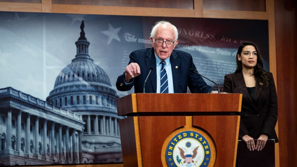 Image Sen. Bernie Sanders speaking at a podium and gesturing with his hand. Rep. Alexandria Ocasio-Cortez is also in the image