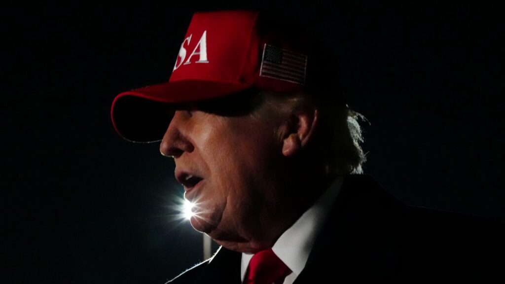 Portrait of Donald Trump at night getting off Air Force One. He's wearing a red hat with white USA lettering