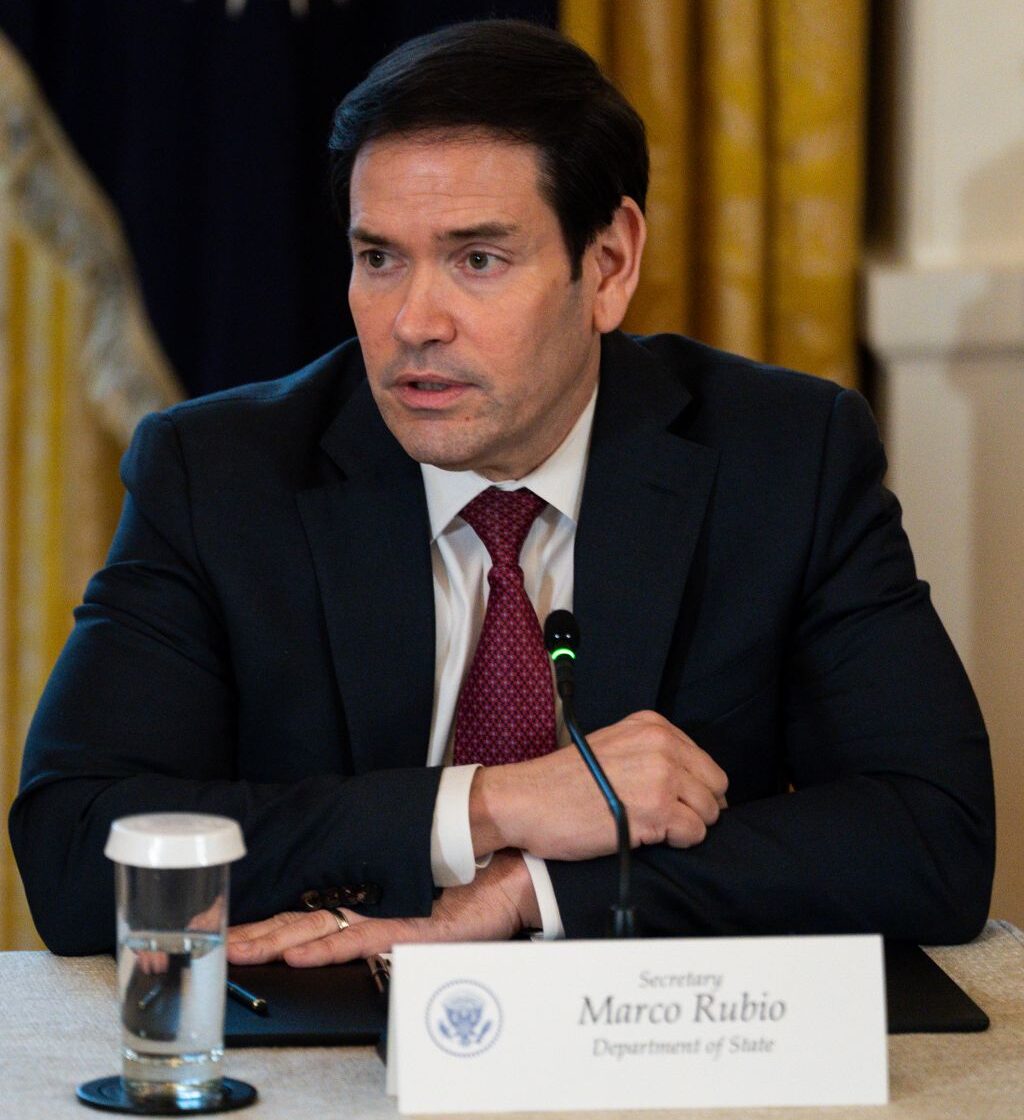 Image of Marco Rubio sitting at a desk with a nametag and a glass of water in front of him