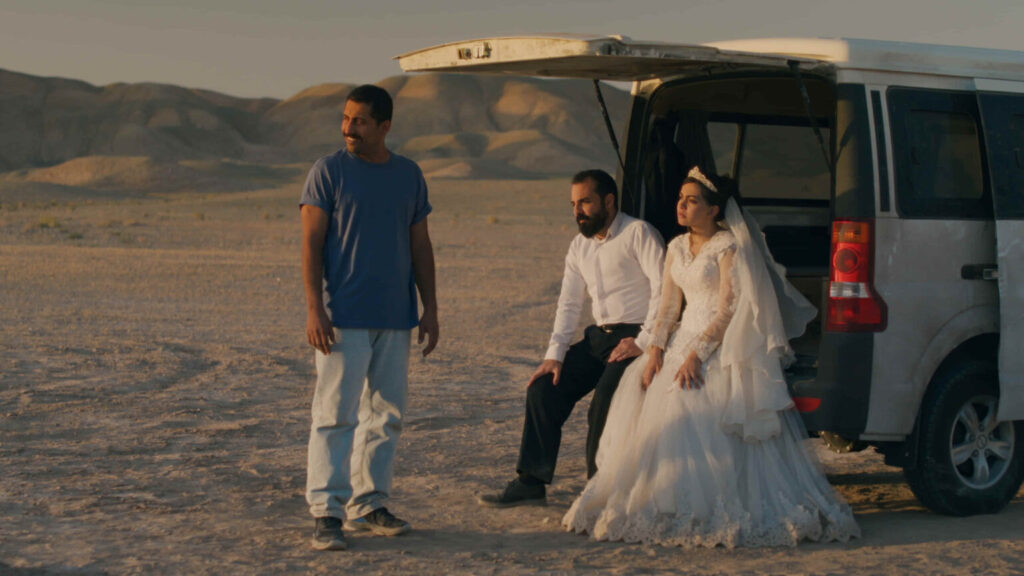 Image of a groom and bride sitting on the tailgate of a pickup truck in the desert alongside a standing man.