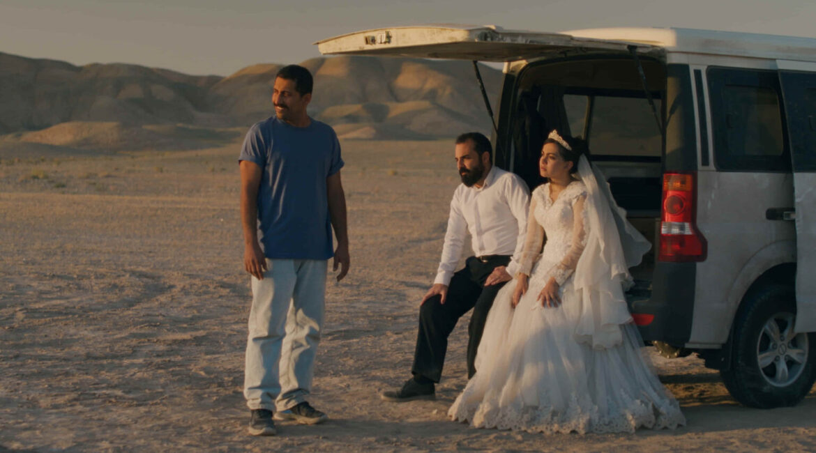 Image of a groom and bride sitting on the tailgate of a pickup truck in the desert alongside a standing man.