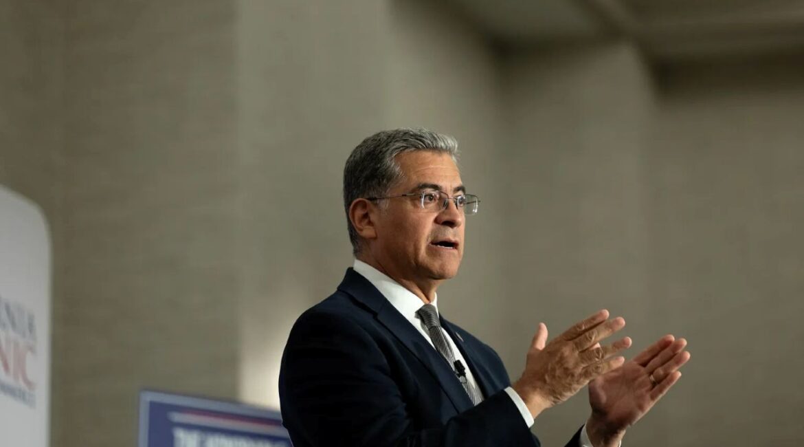 Image of Xavier Becerra in a dark suit with a white shirt, looking left, and gesturing with both hands
