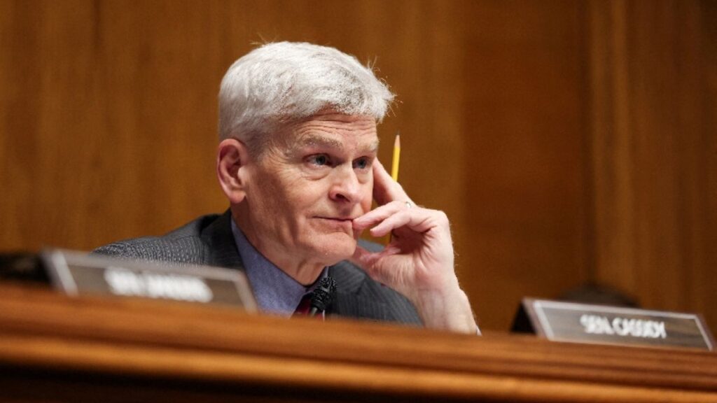 Image of U.S. Senator Bill Cassidy. He's sitting behind a maple-colored wood dais, with his fingers to his face listening