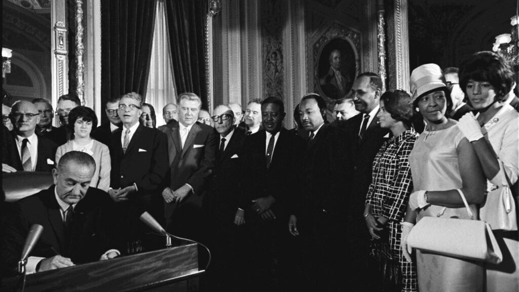 Black and white historic image of LBJ signing the Voting Rights Act as Martin Luther King Jr. and other leaders watch