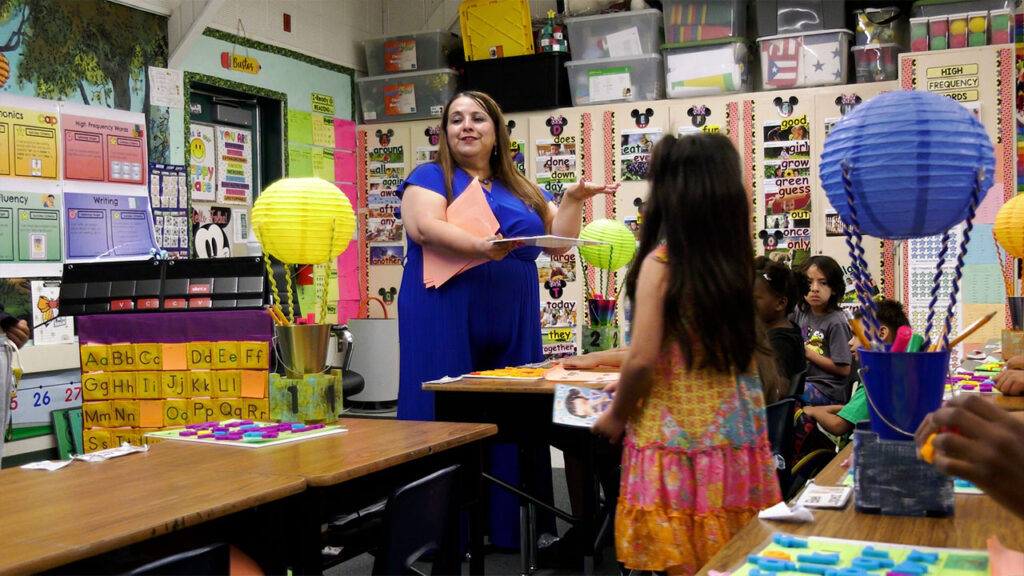Image of teacher Ana Bustos in a royal blue dress holding a folder in a brightly colored classroom as a young female students looks on