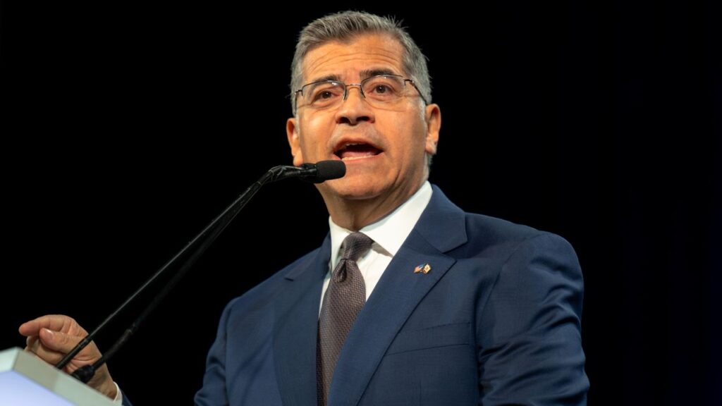 Image of Xavier Becerra speaking at a podium. He's wearing a blue suit, white shirt, and brown tie