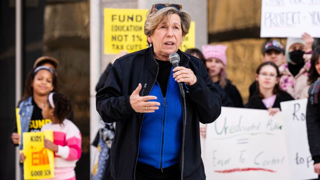 Image of a woman in a blue shirt and dark windbreaker holding a microphone and speaking at a rally