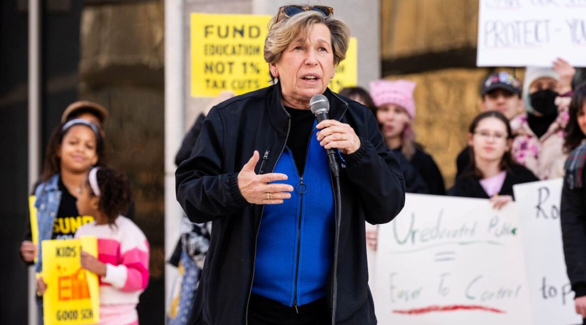 Image of a woman in a blue shirt and dark windbreaker holding a microphone and speaking at a rally
