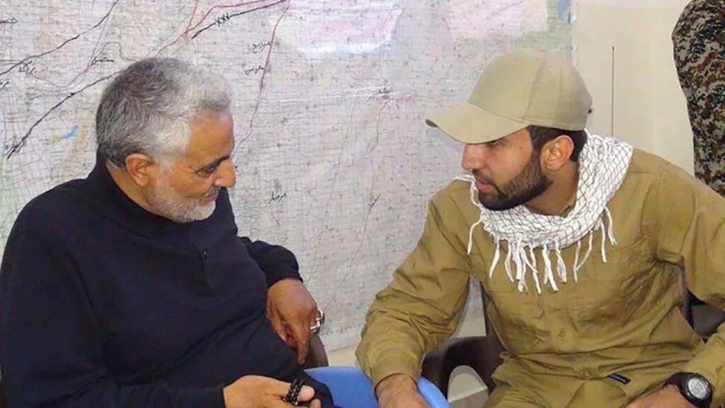 Image of an Iranian general and an Iraqi militia commander facing each other and talking while sitting down in front of a military map