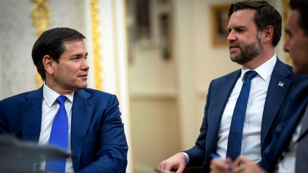 Image of Marco Rubio and JD Vance looking at each other. Both are wearing blue suits, white shirts, and blue ties