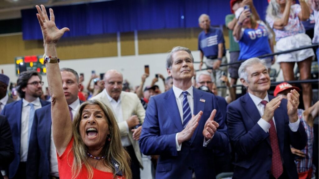 Treasury Secretary Scott Bessent and Dr. Mehmet Oz, right, the administrator of the Centers for Medicare and Medicaid Services, watch and cheer as President Donald Trump speaks during an event at a school in The Villages, Fla., May 1, 2026. The talk show host Dr. Phil McGraw, in a cream jacket, is behind them. (Tom Brenner/The New York Times)