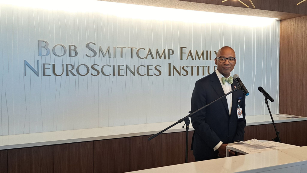 Image of Dr. Ian Johnson, dressed in a dark suit, white shirt, and pastel green bow tie, standing in front of a Bob Smittcamp Family Neorosciences Institute sign inside CRMC