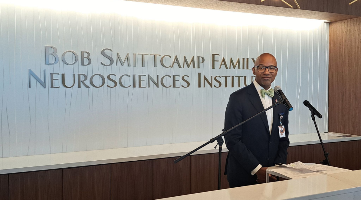 Image of Dr. Ian Johnson, dressed in a dark suit, white shirt, and pastel green bow tie, standing in front of a Bob Smittcamp Family Neorosciences Institute sign inside CRMC