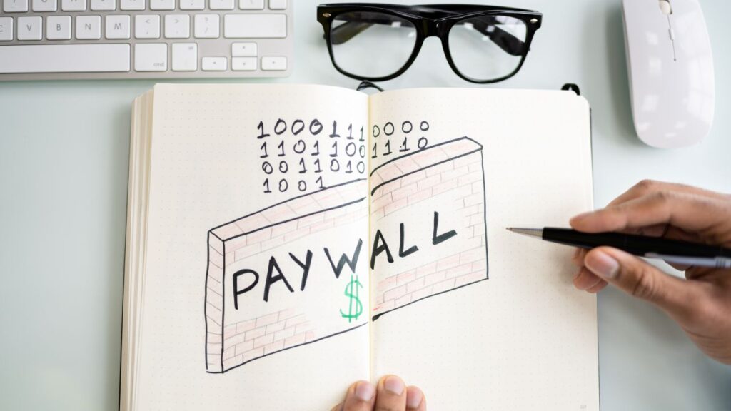Conceptual image of digital news site paywall" a keyboard, black eye glasses, and the word payroll printed on plain white paper that includes computer coding