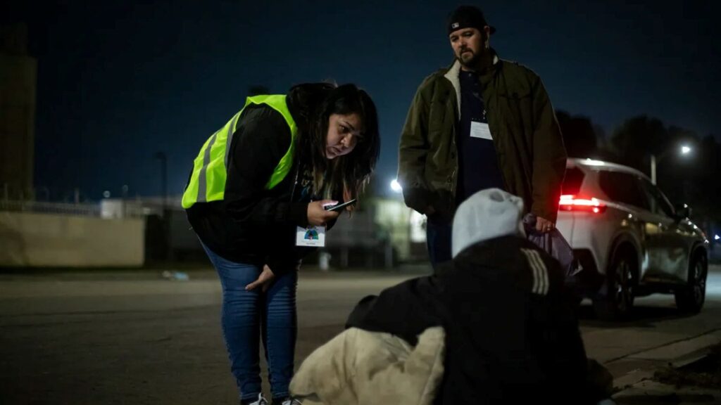 Night image of two people, one in a florescent lime striped safety vest, talking to a homeless person