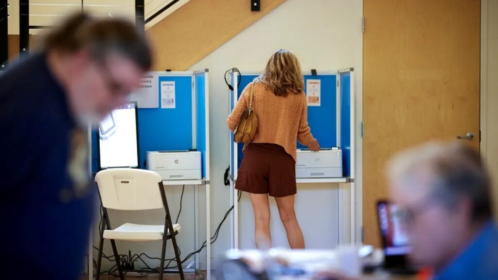 Image of a woman with blonde hair in an orange top and brown shorts filling out a ballot. Her back is to the camera and two men are in the foreground.