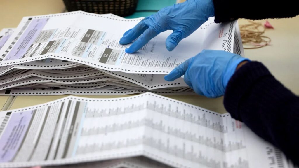 Image of two hands in blue latex gloves going over voting ballots