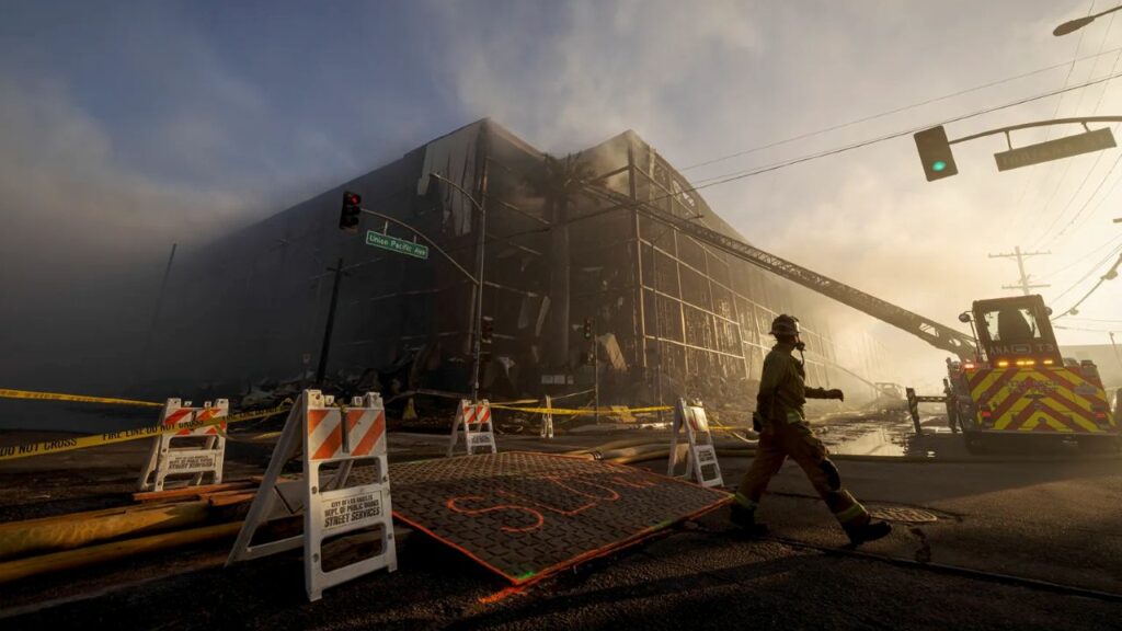 Image of a big building burning with a firefighter and bulldozer on scene, the street closed off, and smoke in the air